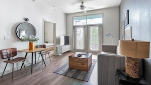 Ein modernes Wohnzimmer mit Couch, Esstisch, runden Stühlen, Fernseher und großem Fenster mit Jalousien sowie einem Holztisch.