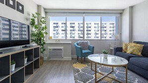 Una sala de estar moderna con televisor de pantalla plana, un sillón azul, un sofá con almohadones amarillos, una mesita y un ventanal con vistas a la ciudad.