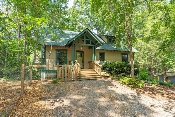 A rustic cabin with a green roof, a wooden porch, and a gravel driveway surrounded by trees. - The Wander Inn | Pet-friendly, Screened Porch, Game Room & Walking Distance to Town! (Black Mountain)