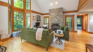 A spacious living room with a stone fireplace, large windows, and a cozy green sofa. - The Wander Inn | Pet-friendly, Screened Porch, Game Room & Walking Distance to Town! (Black Mountain)