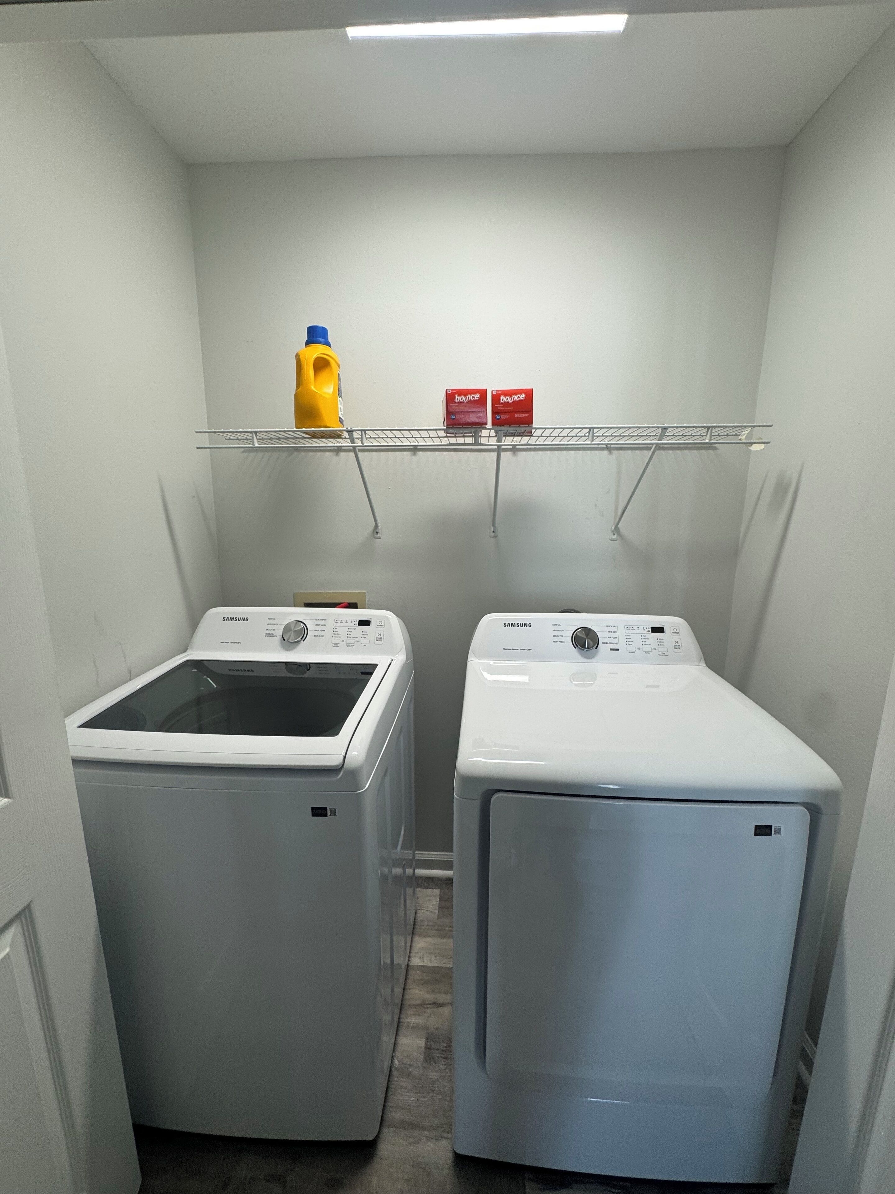 Una sala de bugaderia amb dues rentadores Samsung, una estant amb detergent i una ampolla de detergent groc.