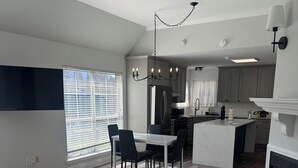 A modern kitchen with a dining area, a fireplace, and a large window with blinds. - Charming 3-bedroom house with AC in Prairieville (Prairieville)