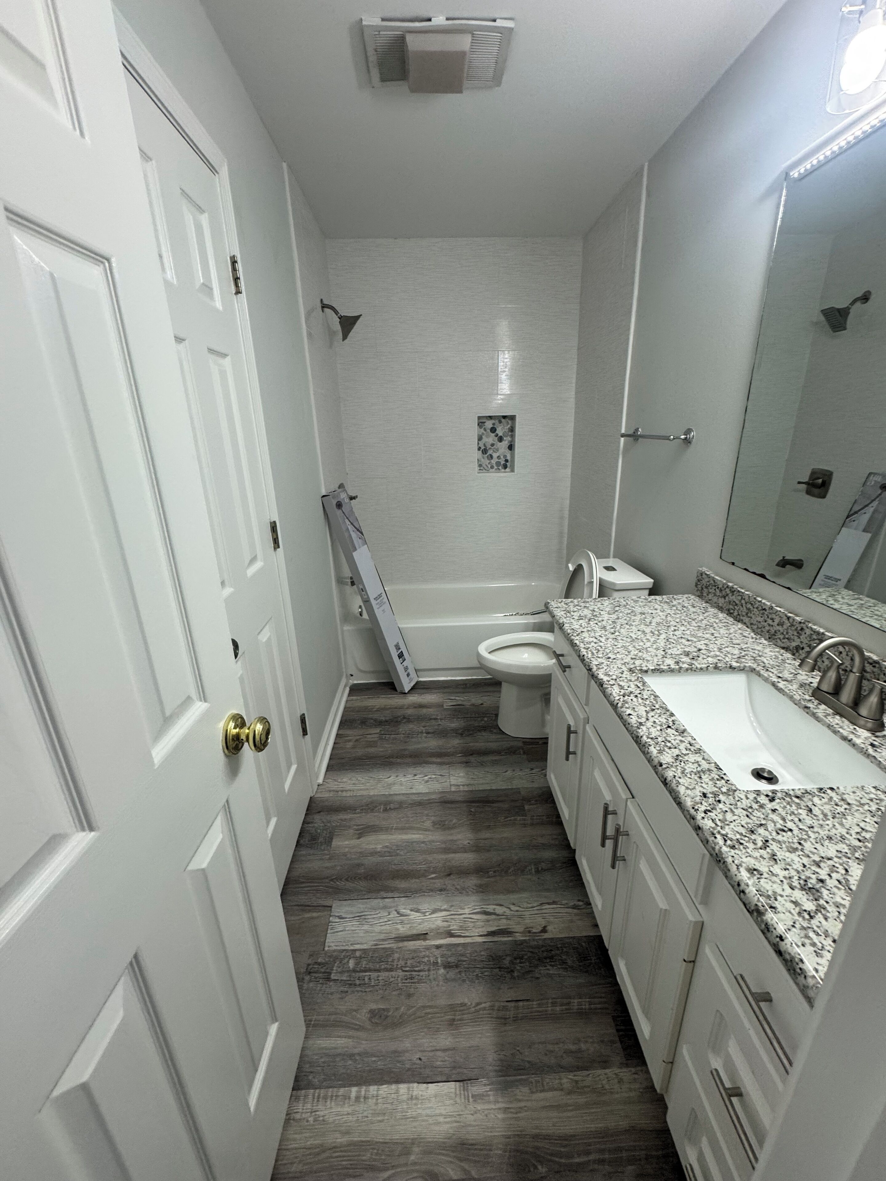 A bathroom with a white door, a shower, a toilet, and a sink with a granite countertop.