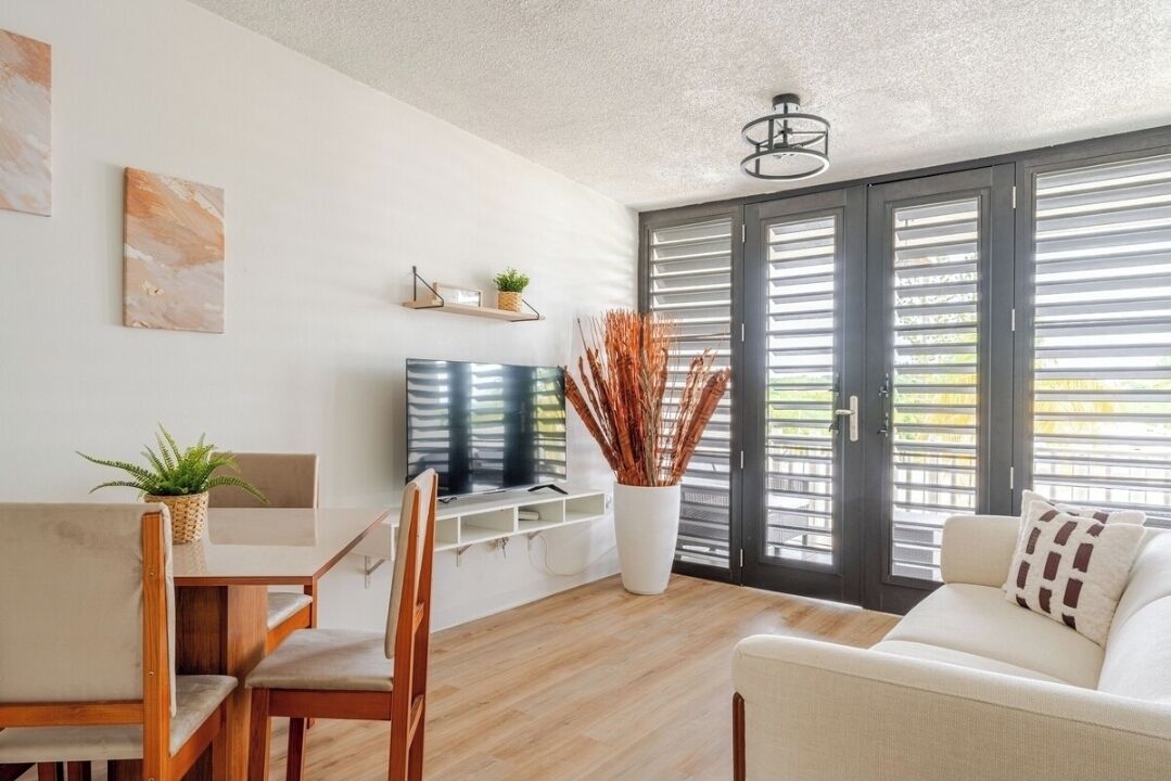 A modern living room with a sofa, a dining table with chairs, a TV, and a large window with blinds.