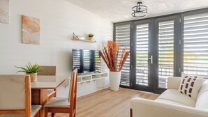 Ein modernes Wohnzimmer mit einer Couch, einem Esstisch mit Stühlen, einem Fernseher und einem großen Fenster mit Jalousien.