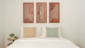 Ein Bett mit weißer Bettwäsche, drei Wandbilder und ein kleiner runder Tisch mit einer Pflanze.