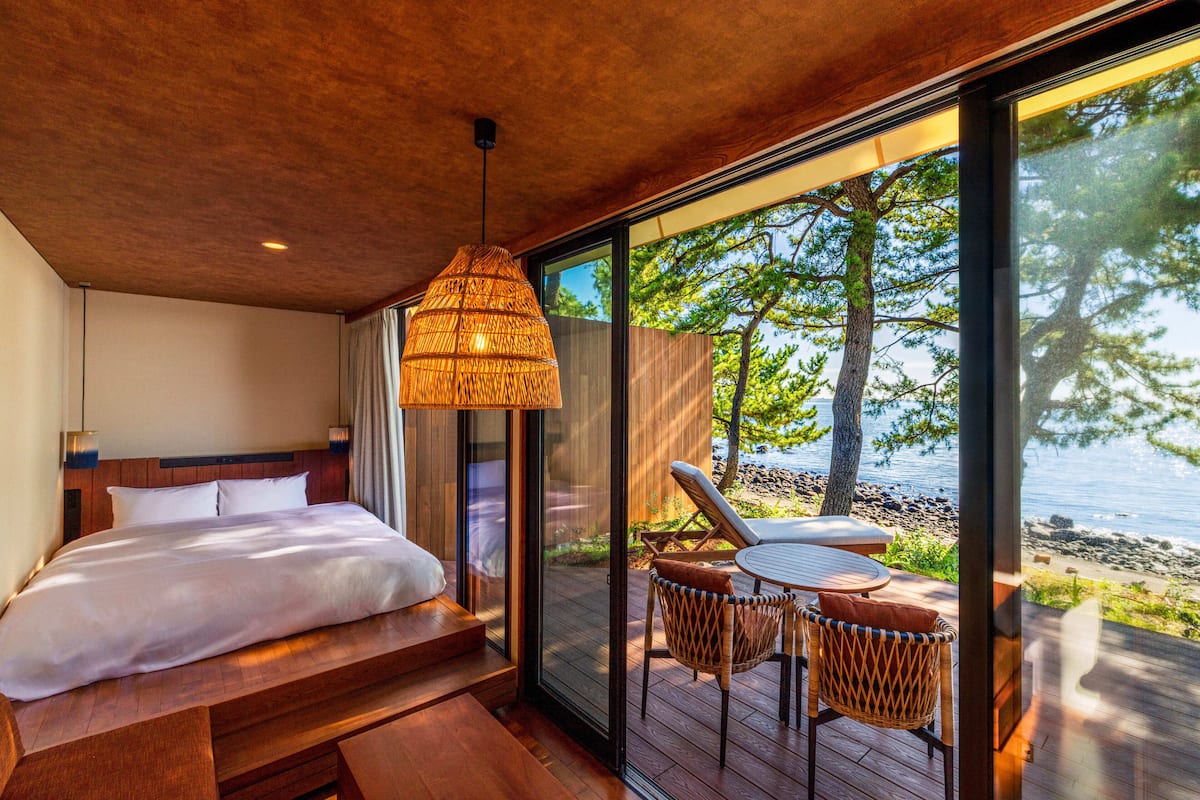 A bedroom with a large bed, a wooden ceiling, and a view of the beach through a glass door.