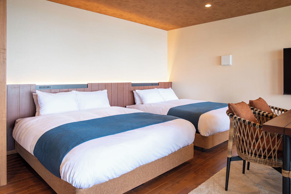 A hotel room with two beds, a wooden floor, a woven chair, and a flat-screen TV mounted on the wall.