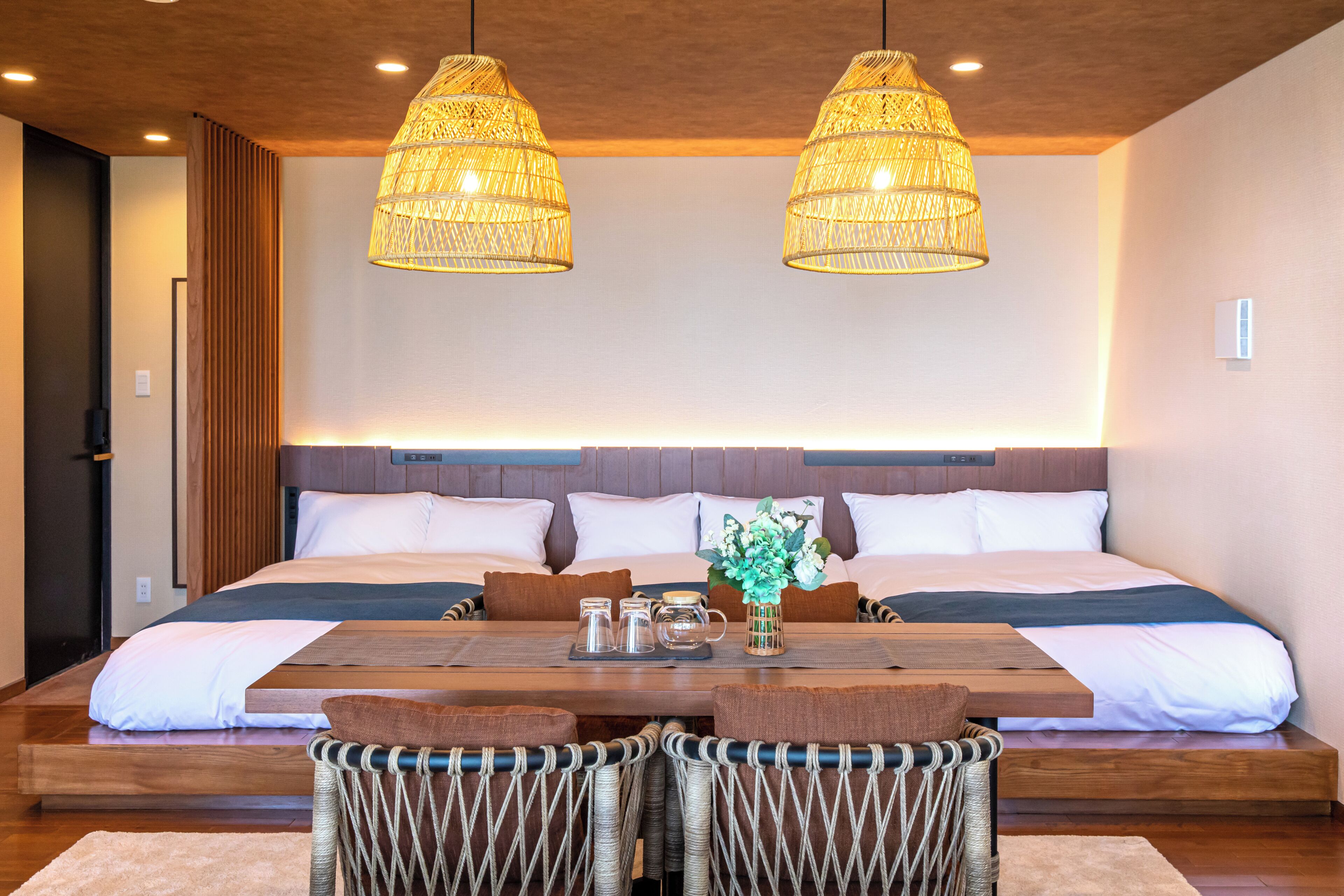 Una habitación de hotel moderna con una cama grande, dos lámparas de mimbre colgantes, una mesa de madera con sillas y un florero con flores.