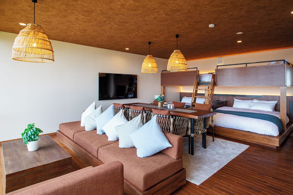 A modern hotel room with a sofa, a dining table, a bunk bed, and hanging wicker lamps.