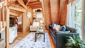 Ein gemütliches Blockhaus mit Holzwänden, einer schwarzen Ledersofa, einem Esstisch und einem großen Fenster mit Blick ins Freie.