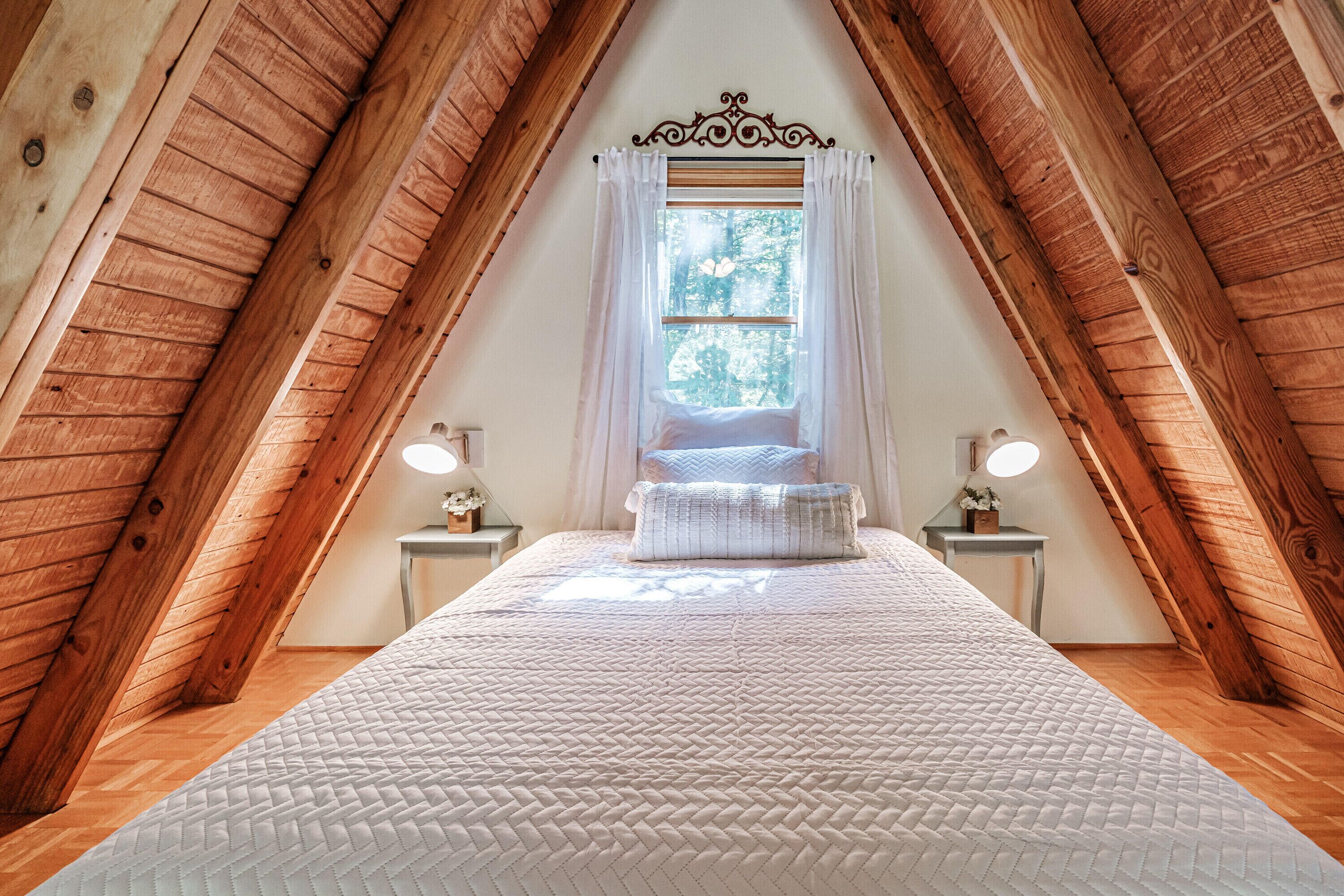 A bedroom with a sloped ceiling, a bed with a white quilted bedspread, two bedside tables with lamps, and a window with sheer curtains.