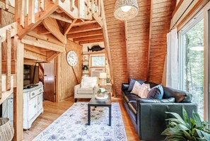 Interior acogedor de una cabaña con paredes de madera, un sofá de cuero negro, una mesa de comedor y un ventanal que ofrece vistas al exterior.