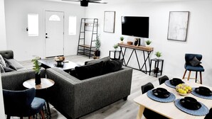 Un salon moderne avec un canapé gris, une table à manger dressée avec des assiettes noires et des sets de table bleus, un téléviseur fixé au mur et un ventilateur de plafond.