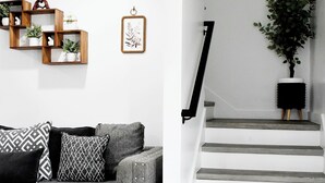 Un salon moderne avec un canapé gris, des coussins à motifs et une étagère en bois agrémentée de plantes. Un escalier avec une rampe noire mène à l’étage supérieur.