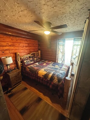Une chambre confortable dans un chalet en rondins, avec un lit en bois, une table de chevet avec une lampe, un ventilateur de plafond et une fenêtre avec des rideaux.