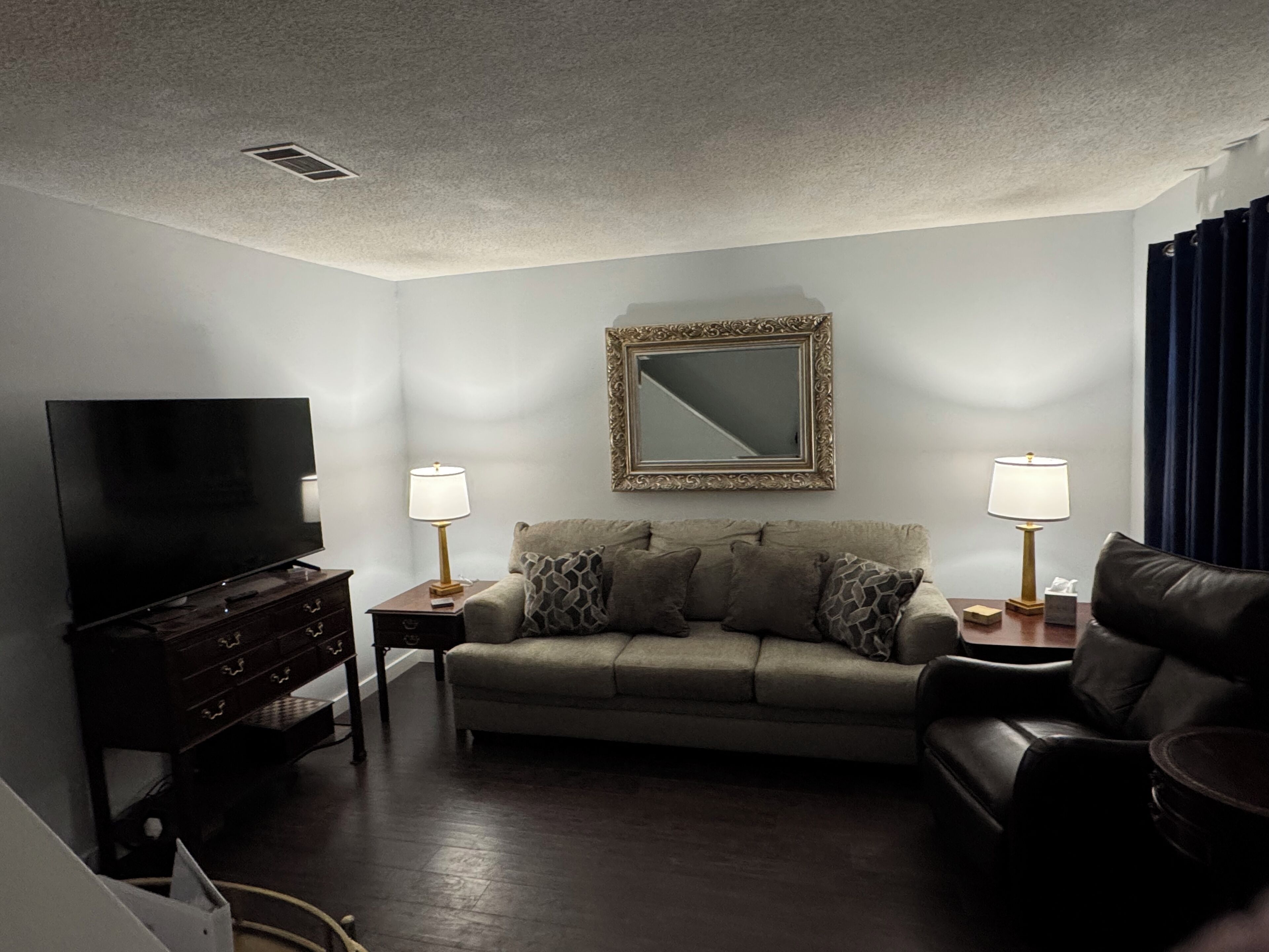 A living room with a flat-screen TV, a sofa, two side tables with lamps, a mirror, and a dark wooden floor.