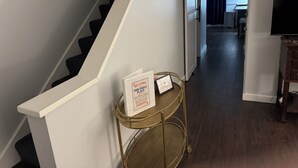 Una escalera con barandilla blanca, una mesita auxiliar con marco dorado y un letrero de bienvenida, y un televisor sobre un soporte de madera en la habitación contigua.