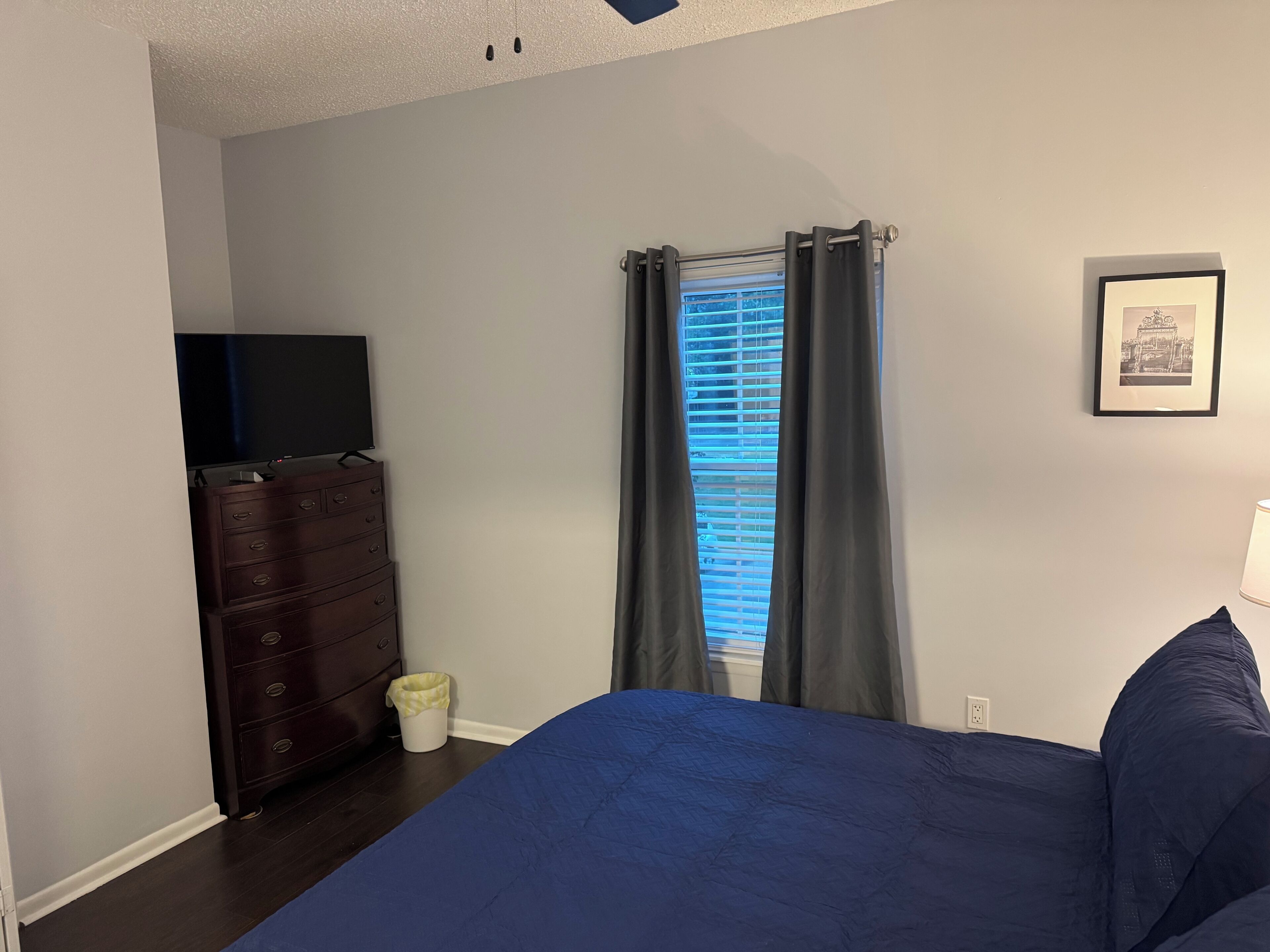 Un dormitorio con cama, cómoda, ventana con cortinas e imagen enmarcada en la pared.