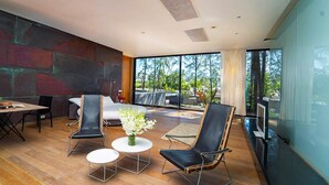A modern living room with a wooden floor, a large window, and a view of trees. There is a dining area with a table and chairs, and a sofa. - At Last You and Your Friends Can Rent a Luxury Villa with 24/7 Concierge (Chang Wat Phang-nga)