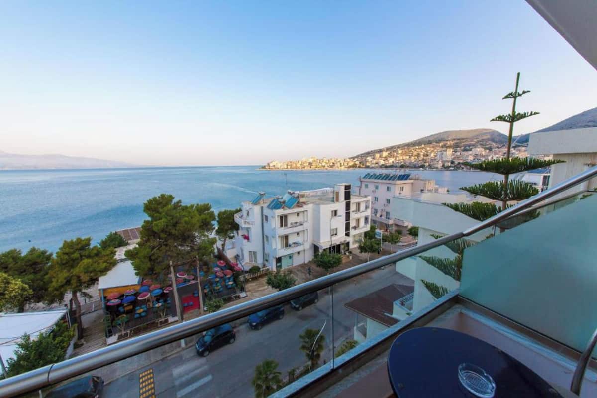 A balcony with a view of the sea, a restaurant with colorful umbrellas, and a building with solar panels.