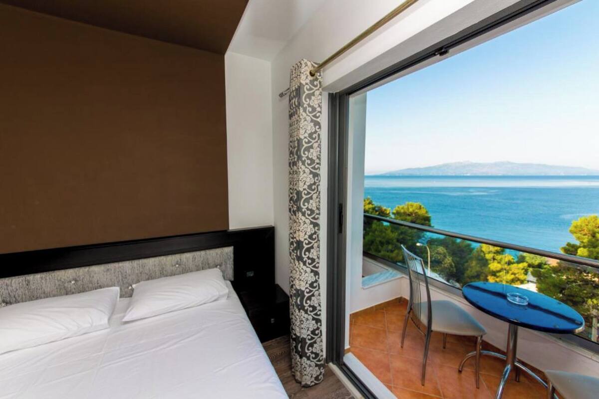 A hotel room with a large bed, a balcony with a table and chairs, and a view of the sea.