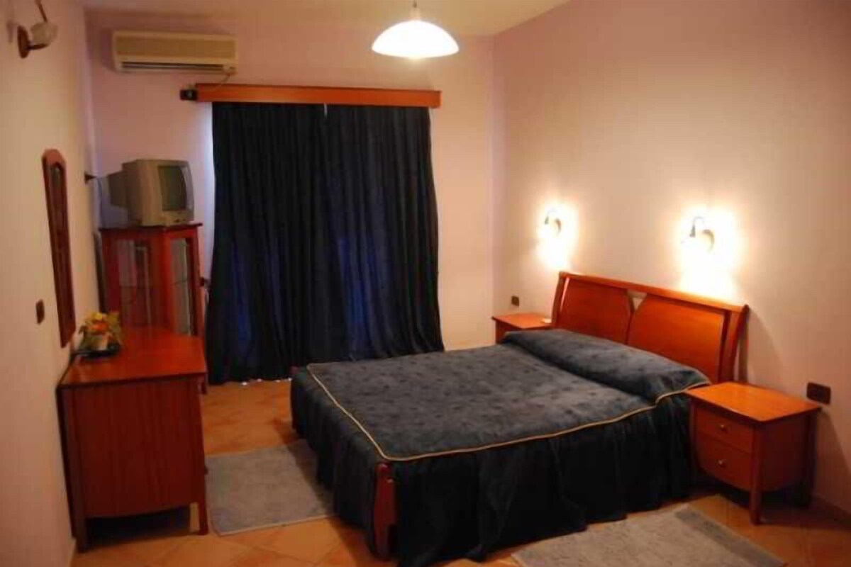 A bedroom with a double bed, two bedside tables, a dresser, a television on a cabinet, and a window with curtains.