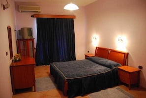 Ein Schlafzimmer mit Doppelbett, zwei Nachttischen, einem Schminktisch und einem Fernseher auf einem Schrank. Ein Fenster mit Vorhängen ist ebenfalls vorhanden.