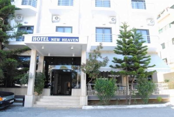 Ένα ξενοδοχείο με το όνομα «Hotel New Heaven», με μια μοντέρνα πρόσοψη, μια σκεπαστή είσοδο και μια μικρή εξωτερική περιοχή με καθίσματα.