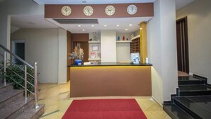 A hotel reception area with a reception desk, multiple clocks, a staircase, and a red carpet. - New Heaven (Sarandë)