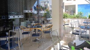 Un espace extérieur aménagé en terrasse, avec des tables et des chaises, un mur en verre et une vue sur un bâtiment en arrière-plan.