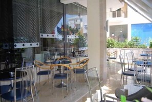 Ένας εξωτερικός χώρος με τραπέζια και καρέκλες, ένας τοίχος από γυαλί και θέα σε ένα κτίριο στο βάθος.