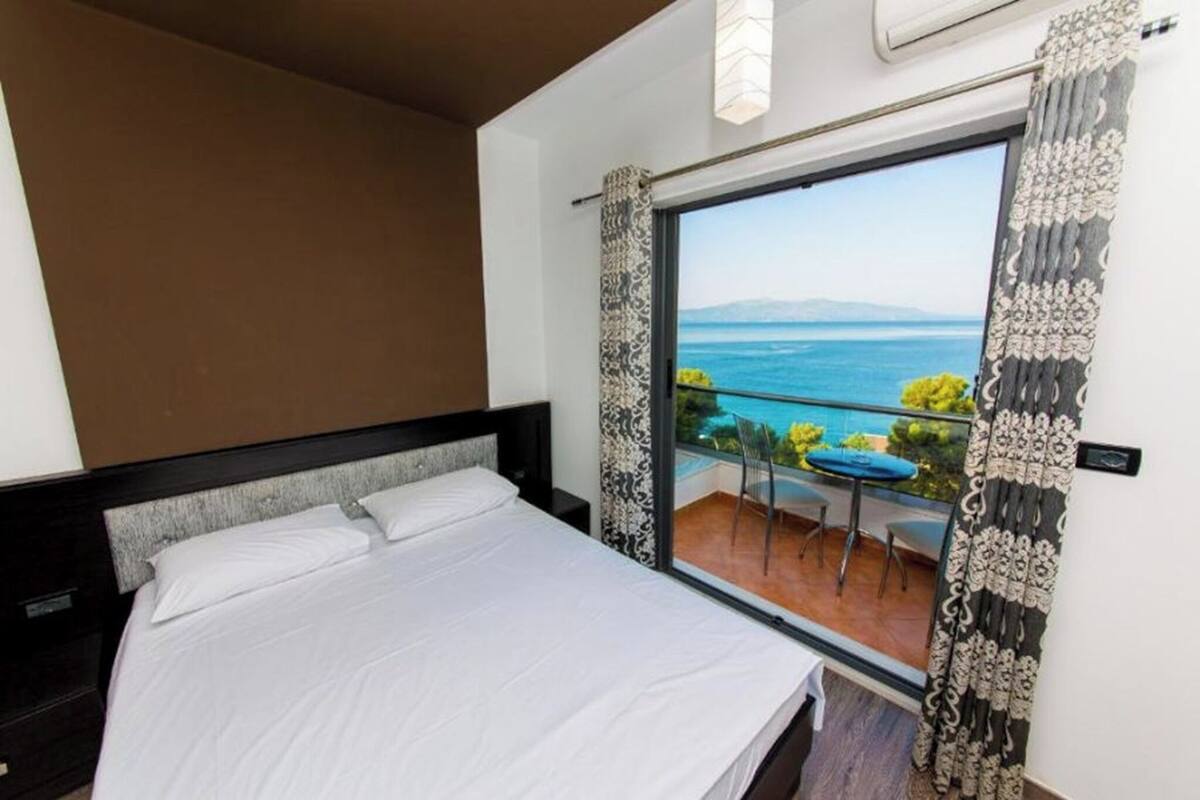 A bedroom with a large bed, a view of the sea through a balcony, and a window with patterned curtains.