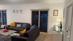 En stue med en mørkebrun sofa, et glastræfabet, og et lille sengebord med en lampe. Der er tre billeder i rammer på væggen, og en skydedør fører ud til en balkon.