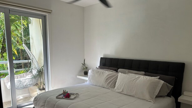 A bedroom with a bed, a bedside table, a hanging chair, and a view of greenery outside.