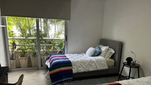 A bedroom with a bed, a small table with a plant, and a view of greenery through a sliding door.