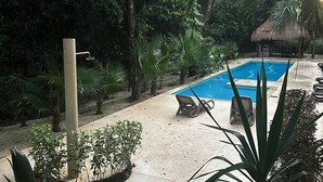 A pool area with lounge chairs and a thatched-roof structure surrounded by dense tropical vegetation.