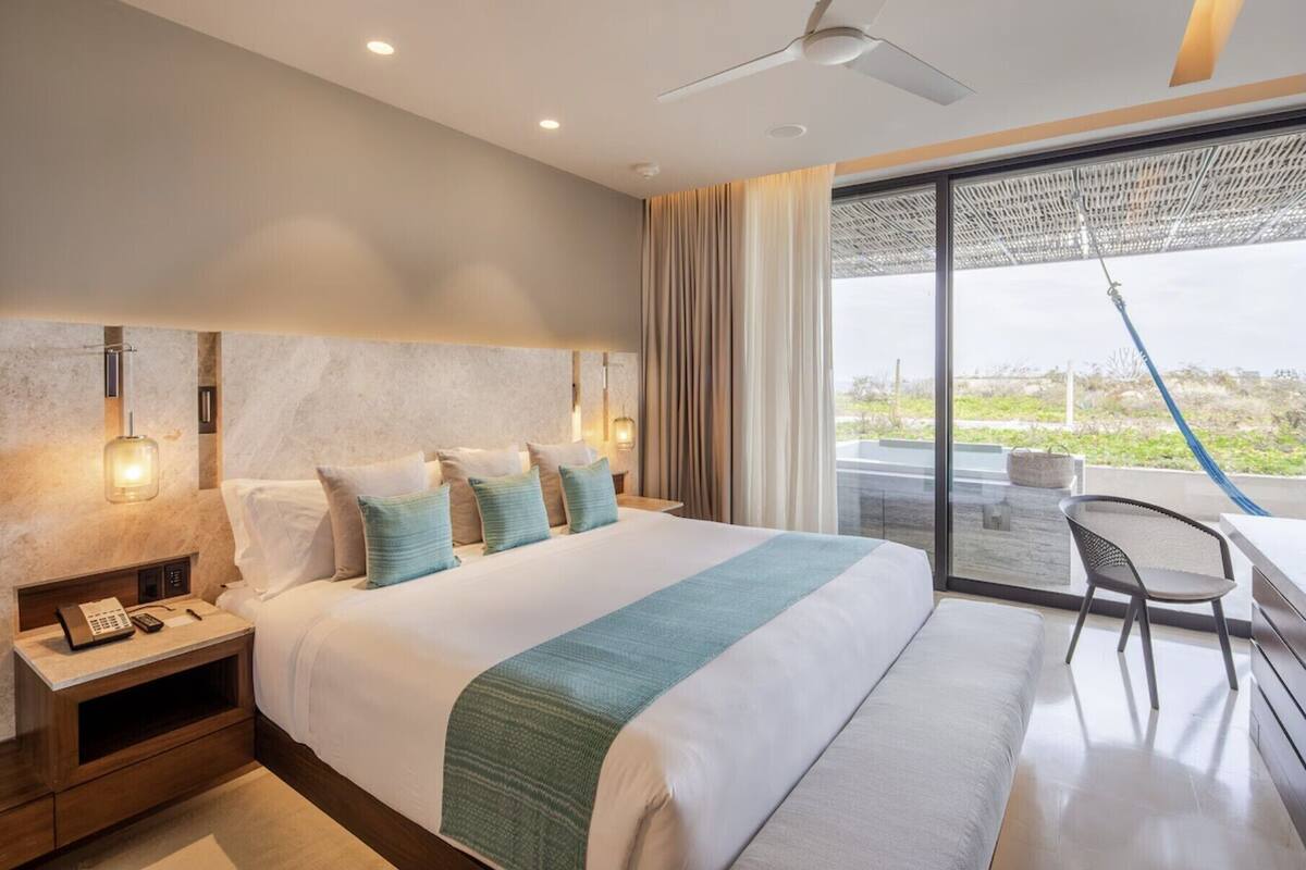 A modern hotel room with a large bed, a desk with a chair, and a view of the outdoors.