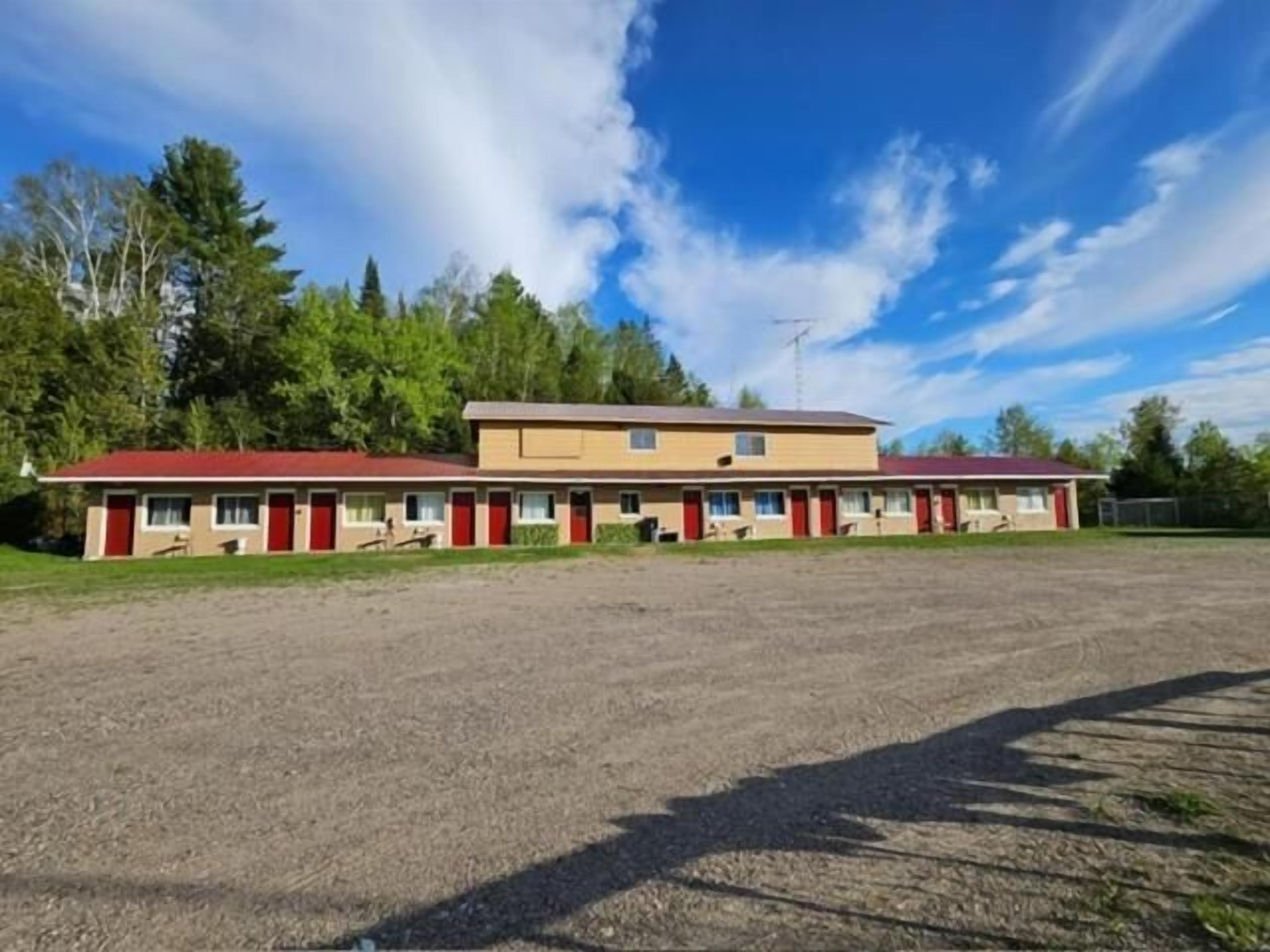 Un motel de dos pisos, con varias habitaciones y puertas rojas, y un estacionamiento al frente.