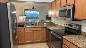 A kitchen with a refrigerator, microwave, oven, granite countertop, wooden cabinets, and a window with a view.