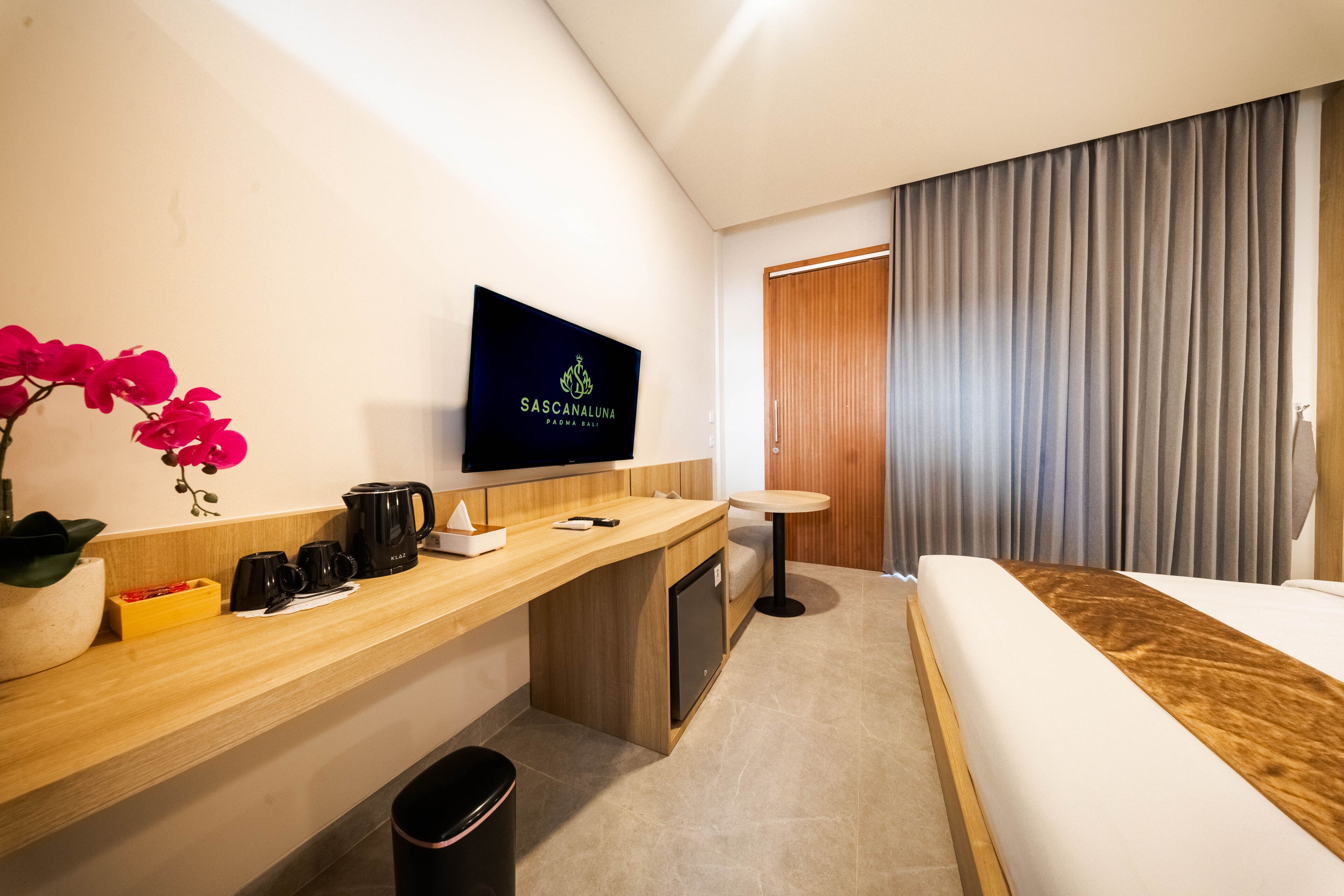 A modern hotel room with a flat-screen TV on the wall, a wooden desk with a coffee maker, a trash bin, and a bed with a wooden headboard.