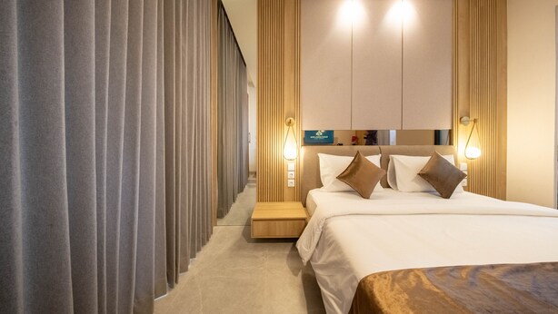 Quarto de hotel moderno com uma cama grande, uma mesa de cabeceira e um criado-mudo. Há luminárias fixadas na parede e uma pequena estante com objetos decorativos.