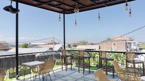 Uma área de estar coberta ao ar livre, com cadeiras e mesas de vime, uma pérgula com luzes penduradas e vista para edifícios próximos.