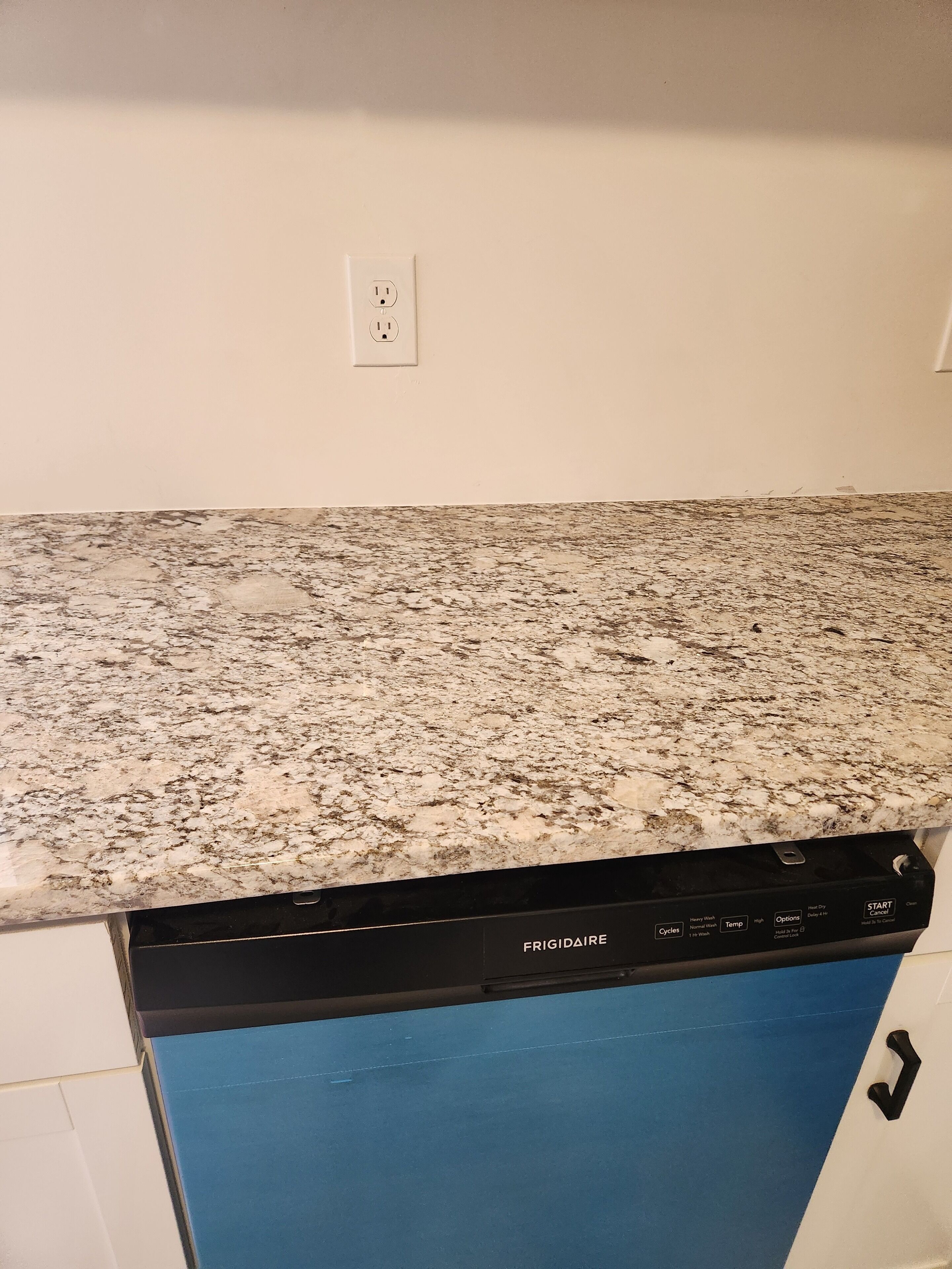A kitchen with a granite countertop, a dishwasher, and a white wall with a power outlet.