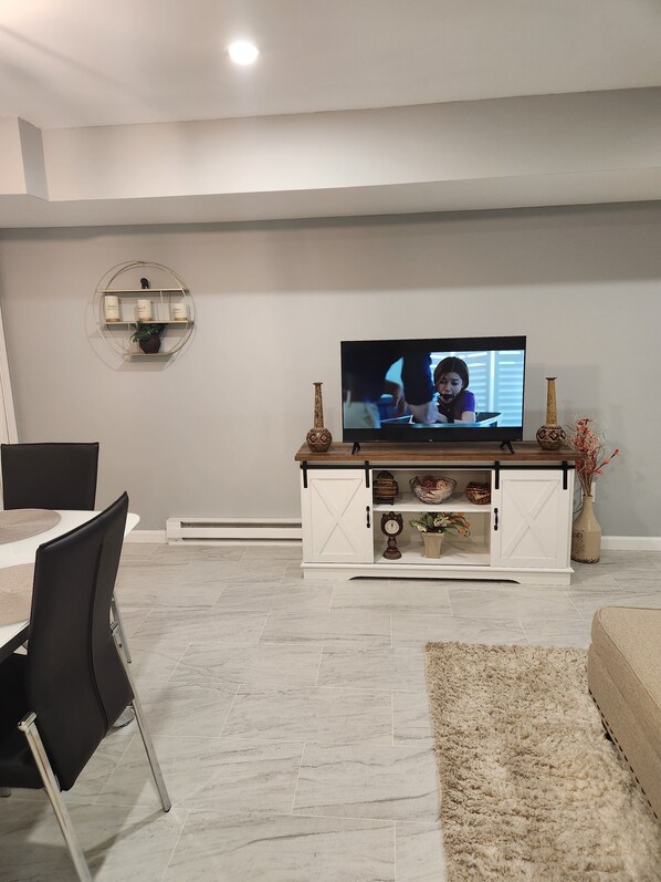 Una sala moderna con televisor de pantalla plana, un comedor con mesa blanca y sillas negras, y un mueble blanco decorado con objetos.