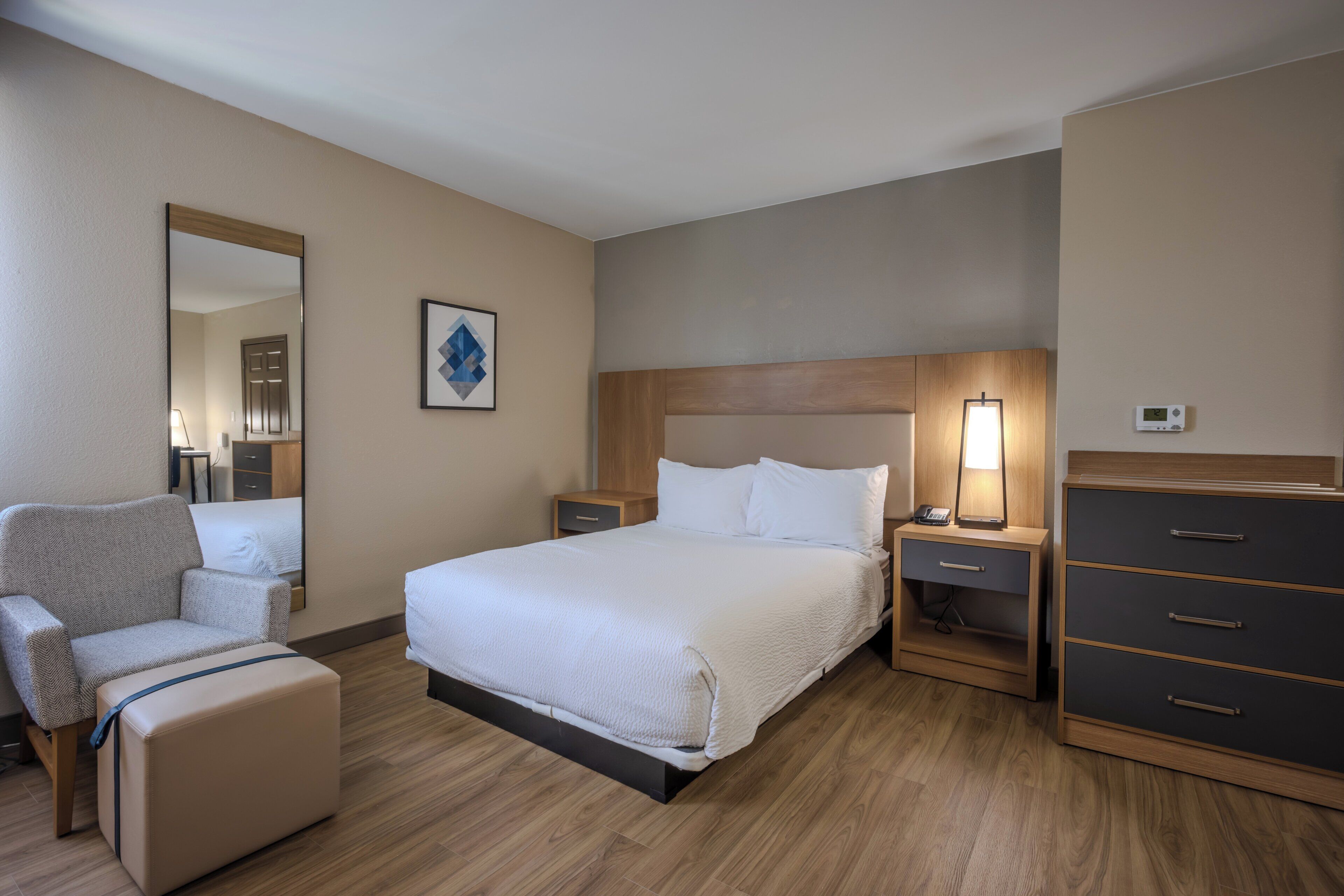 A hotel room with a bed, a chair, a dresser, a nightstand, a lamp, and a thermostat.