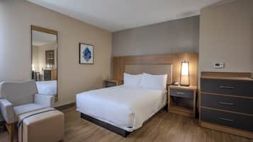 A hotel room with a bed, a chair, a dresser, a nightstand, a lamp, and a thermostat.