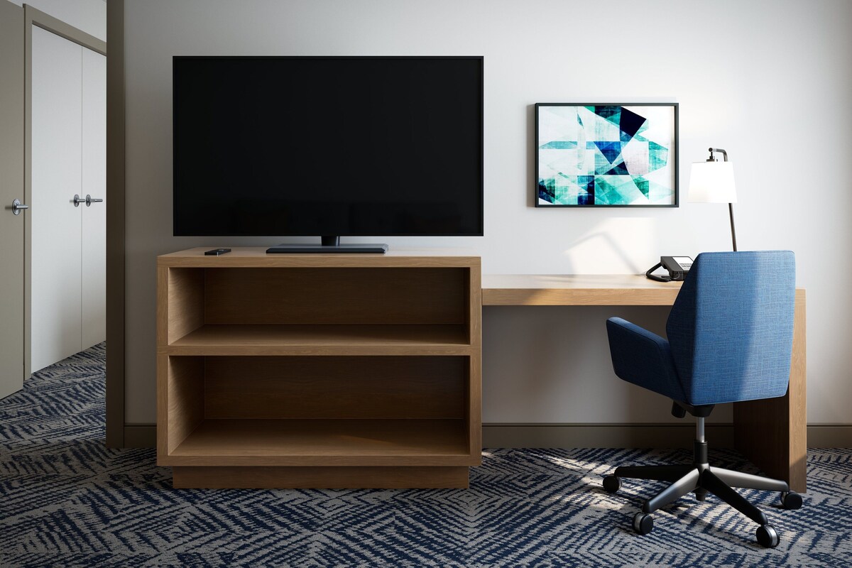 A hotel room with a flat-screen TV, a desk, a chair, and a wall-mounted abstract art piece.