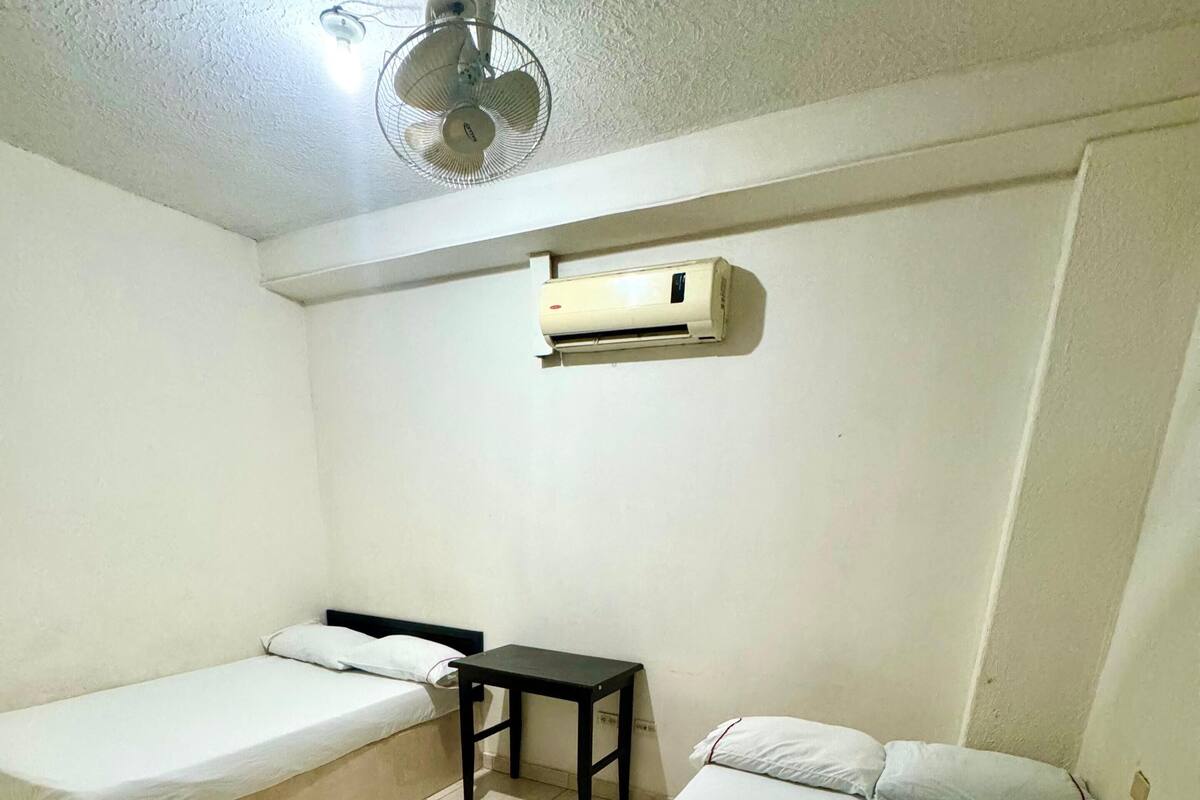 A small, clean room with two single beds, a small table, and a ceiling fan.