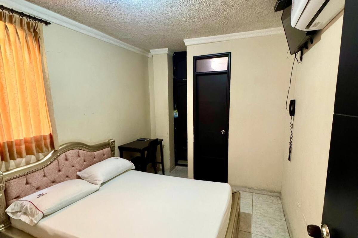 A bedroom with a bed, a desk, a chair, a door, and an air conditioning unit.